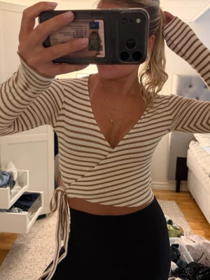 Beige och vit omlottopp med knyt - Säljer en ribbad omlottopp i beige och vitt med smala ränder. Toppen har lång ärm, croppad passform och knyt i sidan. Djup v-ringning framtill och mjukt, stretchigt material som sitter snyggt på kroppen.