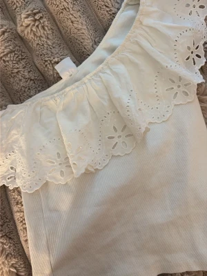 Vit offshoulder topp med spetsvolang - Säljer en söt vit offshoulder topp med bred spetsvolang och broderade blommor. Toppen är ärmlös och har en ribbad struktur i tyget. Perfekt för sommarens alla tillfällen!