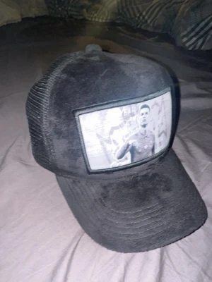 CR7 Caps black - Säljer en svart truckerkeps med mesh på sidorna och ett stort fototryck framtill av en fotbollsspelare. Kepsen har böjd skärm och justerbar snapback baktill. Perfekt för dig som gillar sportig streetstyle.
