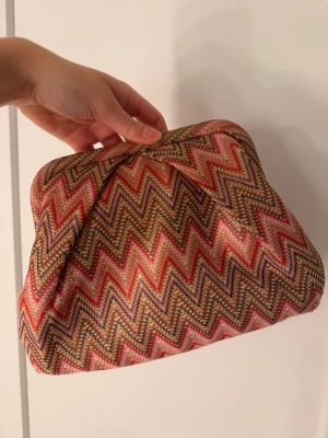 Kuvertväska med zigzag-mönster - Färgglad kuvertväska från Beck Söndergaard med zigzag-mönster i rosa, beige och svart. Väskan har en mjuk form och stängs med metallram. Insidan är svart med en liten ficka med dragkedja. Perfekt för att lyfta din outfit. Säljs inte längre!!🌸