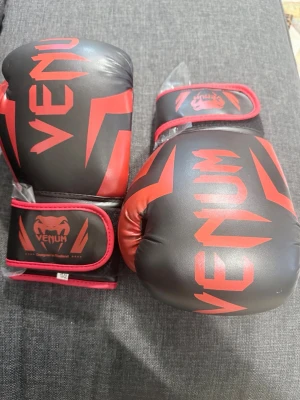 Svarta och röda boxningshandskar Venum - Snygga boxningshandskar från Venum i svart och rött med bred kardborrestängning för extra stöd. Tillverkade i slitstarkt syntetmaterial och har cool design med Venum-logga på ovansidan. Perfekta för träning inom boxning eller kampsport.