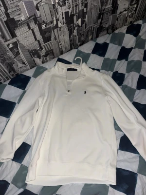 Vit quarter zip från Polo Ralph Lauren - Klassisk vit långärmad quarter zip från Polo Ralph Lauren med liten broderad logga på bröstet. Tröjan har hög krage med dragkedja och är tillverkad i mjukt material som känns skönt mot huden. Perfekt till jeans eller chinos.
