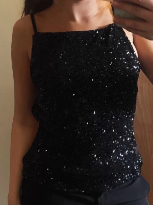 Svart glittrig paljettopp med öppen rygg - Snygg svart topp med paljetter och tunna axelband. Toppen har en glittrig finish och öppen rygg med smala band som ger en trendig look. Perfekt för dig som vill sticka ut och glänsa lite extra.