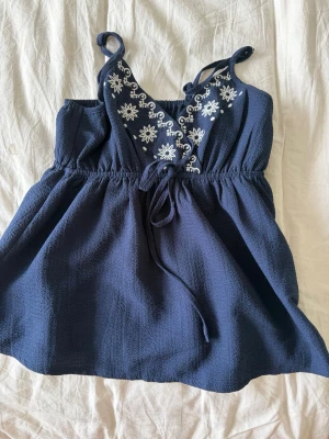 Mörkblå broderad linnetopp - Supersöt mörkblå linnetopp med vita broderade blommor framtill och knyt i midjan. Toppen har smala axelband och en luftig passform, perfekt för varma dagar. Materialet känns lätt och svalt mot huden.