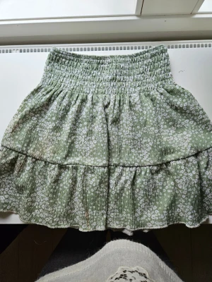 Blommig kjol - Söt grön kortkjol från Gina Tricot med vitt blommönster och smock i midjan. Kjolen har volangdetaljer och är gjord i ett lätt och luftigt material, perfekt för varma dagar.