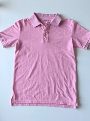 Rosa pikétröja från GAP - Säljer en klassisk rosa pikétröja från GAP med korta ärmar och krage. Perfekt för en avslappnad och stilren look. Är egenligen barnstorlek, men sitter som en XS man.