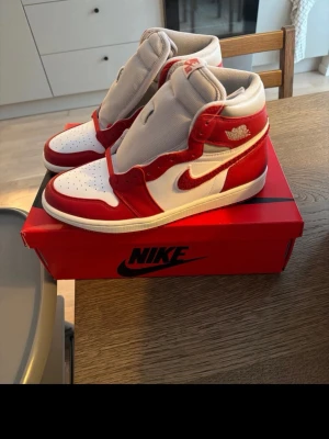 Nike Air Jordan 1 High röd/vit - Säljer ett par Nike Air Jordan 1 High sneakers i rött och vitt med klassisk Swoosh och Air Jordan-logga på sidan. Skorna har högt skaft, perforerad tåbox och kommer med extra vita skosnören. Materialet är en mix av läder och textil för en ikonisk look.