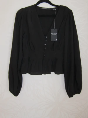 Svart blus med knappar och v-ringning - Stilren svart blus med v-ringning och knappar framtill. Blusen har långa puffärmar med resår vid ärmslut och en lätt volang nertill. Perfekt för en trendig och elegant look. Materialet ser ut att vara lätt och luftigt.
