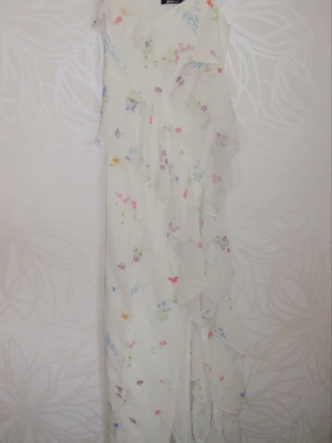 Lång blommig klänning med volanger - Romantisk långklänning i ljus färg med blommigt mönster och volangdetaljer längs sidan. Klänningen har ärmlös design och ett luftigt, svepande fall som ger en drömmig känsla. Perfekt för sommarens alla tillfällen.