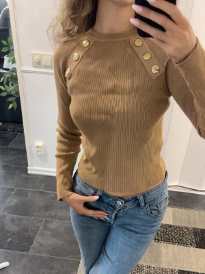 Ribbad brun tröja med guldknappar - Snygg ribbad tröja i brun färg med långa ärmar och dekorativa guldknappar på axlarna. Tröjan har en kroppsnära passform och rund halsringning. Perfekt att matcha med jeans för en trendig look.