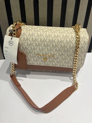 Beige och brun Michael Kors handväska - Snygg handväska från Michael Kors i beige canvas med MK-logga och bruna läderdetaljer. Väskan har guldfärgad kedjerem och präglad logotyp framtill. Perfekt storlek för det viktigaste och ger en lyxig touch till din outfit.