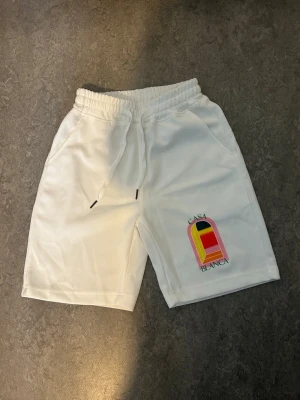 Vita shorts med tryck och snörning - Säljer ett par vita shorts med färgglatt tryck på ena benet och texten 'Casa Blanca'. Shortsen har elastisk midja med snörning och sidofickor. Perfekta för sommaren och har en avslappnad passform.