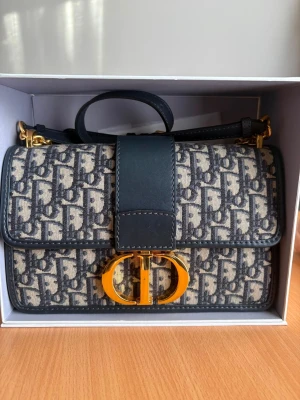 Dior handväska med monogram och guldspänne - Snygg handväska från Dior med klassiskt blått och beige monogrammönster, guldfärgat CD-spänne och detaljer i mörkblått läder. Väskan har både handtag och axelrem samt exklusiv känsla med gulddetaljer. Perfekt för dig som gillar lyxiga accessoarer.