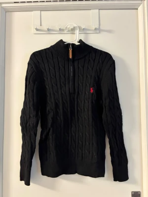 Svart kabelstickad tröja Polo Ralph Lauren - Kabelstickad svart tröja från Polo Ralph Lauren med halv dragkedja och röd broderad logga på bröstet. Tröjan har hög krage och långa ärmar, perfekt för kyliga dagar. Klassisk och tidlös design som passar till det mesta.
