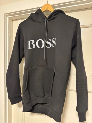Svart hoodie med BOSS-tryck - Snygg svart hoodie med stor vit BOSS-text på bröstet. Klassisk modell med huva och dragsnören samt en stor känguruficka framtill. Tillverkad i mjukt material som passar perfekt för en avslappnad stil. TVÄTTAS INNAN!