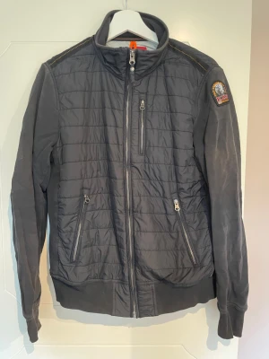 Svart quiltad jacka Parajumpers, storlek S - Stilren svart quiltad jacka från Parajumpers med ståkrage, dragkedja framtill och två snedställda fickor med dragkedja. Jackan har ribbade muddar och ett märke på ena ärmen.