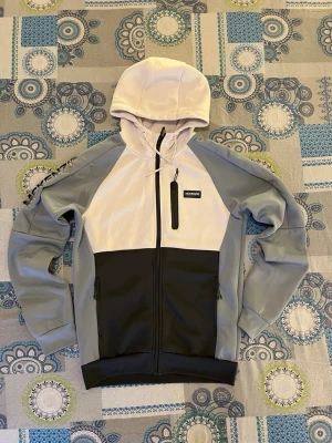 Grå och vit hoodie från McKenzie - Snygg hoodie från McKenzie i färgerna grå, vit och svart. Modellen har dragkedja framtill, två sidofickor med dragkedja och en bröstficka. Tillverkad i mjukt material med huva och justerbar dragsko. Perfekt för en avslappnad streetwear-look.