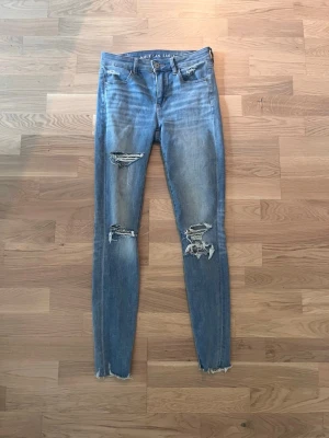 Blå slitna skinny jeans - Säljer ett par blå skinny jeans från American Eagle med slitningar och hål på benen för en trendig look. Jeansen har hög midja, klassisk femficksmodell och råa kanter vid bensluten. Perfekta för dig som gillar en avslappnad och edgy stil. Storlek 4.