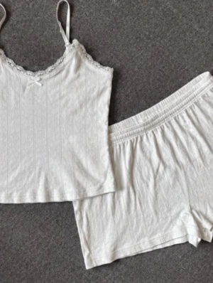 Vitt spetskantat linne och shorts-set - Sött set med vitt linne och matchande shorts i mjukt tyg. Linnet har smala axelband, spetskant vid urringningen och en liten rosett framtill. Båda delarna har diskret hålmönster och känns riktigt bekväma. Perfekt för dig som gillar en enkel men gullig stil.