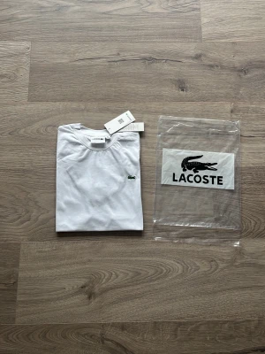 Vit t-shirt från Lacoste - Klassisk vit t-shirt från Lacoste med den ikoniska gröna krokodilloggan broderad på bröstet. T-shirten har rund hals, korta ärmar och är tillverkad i mjuk bomull. Perfekt basic-plagg för en clean och stilren look.