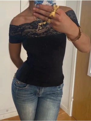 Svart offshoulder topp med spets - Snygg svart offshoulder topp med spetsdetaljer över bysten och axlarna. Toppen är tight och har korta ärmar. Perfekt för dig som vill ha en trendig och feminin look med lite extra edge.