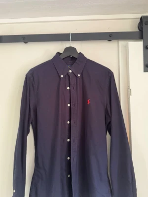 Mörkblå skjorta från Ralph lauren - Snygg mörkblå skjorta med klassisk krage och vita knappar hela vägen framtill. Skjortan har lång ärm och en liten röd broderad logga på bröstet. Perfekt för dig som gillar stilrena plagg med en enkel men elegant look.