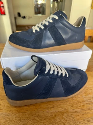 Maison margiela gats - Snygga mörkblå sneakers med kontrasterande mockadetaljer och vita snören. Skorna har en klassisk låg profil och en brun gummisula som ger en retrovibe. Ovandelen kombinerar slätt läder och mocka för en stilren look. Perfekta för dig som gillar minimalistisk och tidlös design.