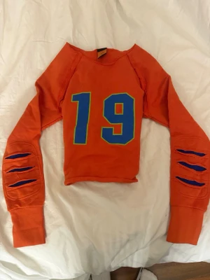 Orange croppad topp Harley-Davidson - Cool orange croppad långärmad topp från Harley-Davidson med blått och gult tryck av siffran 19 på bröstet. Ärmarna har unika slitsade detaljer med blått tyg under. Perfekt för dig som vill sticka ut med en sportig vibe. Passar xxs-m.