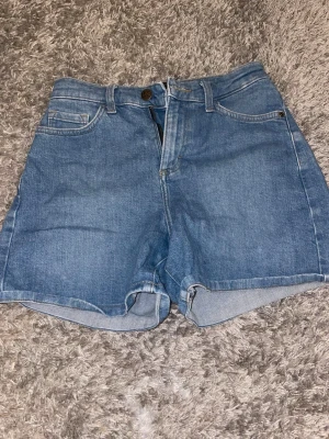 Blå denimshorts från Lee - Klassiska blå jeansshorts från Lee med medel midja och femficksmodell. Tillverkade i slitstark bomullsdenim med knapp och dragkedja framtill. Perfekta för sommaren och har en avslappnad passform.