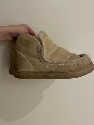 Beiga MOUs - Säljer ett par beiga moon boots med mjuk mockaliknande yta och stickade detaljer runt kanten. Skorna har en vadderad ovandel och grov sula, perfekta för kalla dagar. De har en unik design med synliga sömmar och mysig känsla.