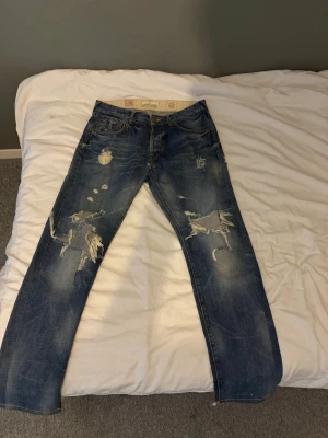 Blå slitna jeans med raka ben - Säljer ett par blå jeans med raka ben och tydliga slitningar samt hål på både fram- och baksidan. Jeansen har klassisk femficksdesign och en tvättad look som ger en cool, avslappnad stil. Perfekta för dig som gillar streetwear och en trendig vibe.