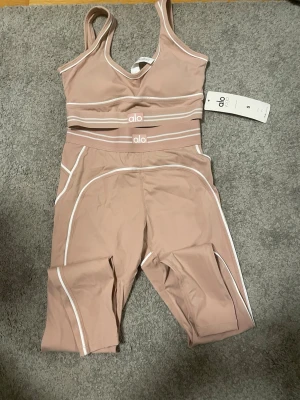 Rosa set från Alo Yoga - Snygg beige sport-bh från Alo Yoga med vita detaljer och bred resår under bysten. Bh:n har en mjuk och stretchig passform med vadderade kupor och är perfekt för yoga eller träning. Materialet är slätt och bekvämt mot huden. Byxorna kommer även med.