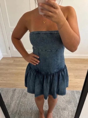 Ärmlös jeansklänning med volang - Snygg ärmlös jeansklänning i blå denim med volangkjol och smockad rygg. Axelbandslös modell som sitter tajt upptill och har en utställd kjol. Perfekt för varma dagar och enkel att styla med accessoarer.