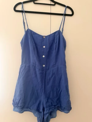 Blå jumpsuit med knappar framtill - Snygg blå jumpsuit med tunna axelband och fyra knappar framtill. Kort modell med avslappnad passform och lätt, luftigt tyg som känns perfekt för varma dagar. Enkel och stilren design som är lätt att matcha med accessoarer.