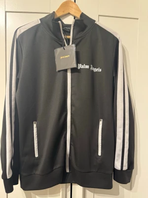 Palm Angels svart track jacket - Svart zip up från Palm Angels med vita ränder längs ärmarna och vit dragkedja. Den är i storlek M och den passar som M/S den är i bra väldigt bra skick. 