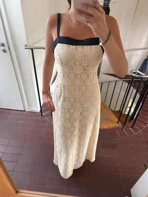 Lång spetsklänning med svarta band - Elegant långklänning i beige spets med svarta axelband och kant upptill. Klänningen har en rak passform och är ärmlös med fina spetsdetaljer över hela plagget. Perfekt för dig som vill ha en stilren och romantisk look.