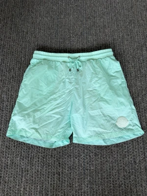 Mintgröna badbyxor från Moncler - Snygga mintgröna badbyxor från Moncler med elastisk midja och dragsko. Tillverkade i ett lätt och snabbtorkande material. Moncler-logga i vitt på vänster ben ger en clean och exklusiv look. Perfekta för stranden eller poolen.