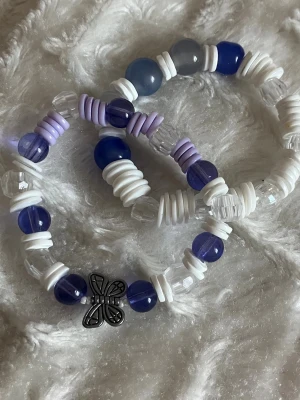 Handgjorda pärlarmband med fjäril - Två elastiska armband med pärlor i lila, vitt, blått och transparent. Ena armbandet har en metallfjäril som detalj. Pärlorna är både runda och platta 😊
