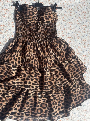 Leopardmönstrad volangklänning - Säljer en axelbandslös kort klänning med leopardmönster och volanger. Klänningen har smockad överdel som ger en snygg passform och ett flowy fall. Perfekt för dig som vill sticka ut med ett djuriskt print och cool look.