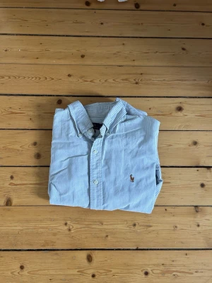 Randig skjorta från Polo Ralph Lauren - Säljer en klassisk blåvit randig skjorta från Polo Ralph Lauren i slim fit-modell. Skjortan har knappar framtill, button-down krage och broderad logga på bröstet. Perfekt för en stilren look.