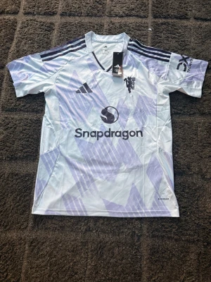 Manchester United bortatröja Slim fit - Säljer en Manchester United bortatröja från Adidas i storlek S slim fit. Tröjan är vit med ljuslila geometriskt mönster, har mörklila Adidas-ränder på axlarna och klubbmärke på bröstet. Tillverkad i lätt och ventilerande material, perfekt för fotboll eller träning.