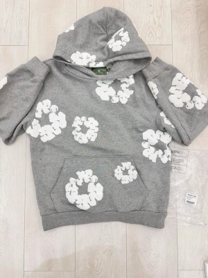 Denim tears  hoodie  - Snygg grå hoodie med korta ärmar och stora vita blommönster över hela tröjan. Har en klassisk huva och en stor magficka framtill. Perfekt för dig som vill sticka ut med en unik och trendig stil. Materialet ser ut att vara mjukt och bekvämt, troligen bomullsblandning.