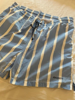 Blåvitrandiga badbyxor från Hackett - Snygga badbyxor från Hackett i ljusblått med vita vertikala ränder. De har elastisk midja med svart dragsko och en bakficka med lock. Perfekta för stranden eller poolen, klassisk design som sticker ut.