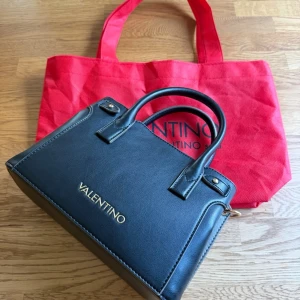 Svart handväska från Mario Valentino - Snygg svart läderhandväska från Mario Valentino  med guldfärgade detaljer och logga framtill. Väskan har en avtagbar axelrem. Kommer med en röd tygpåse med Mario Valentino-tryck. 
