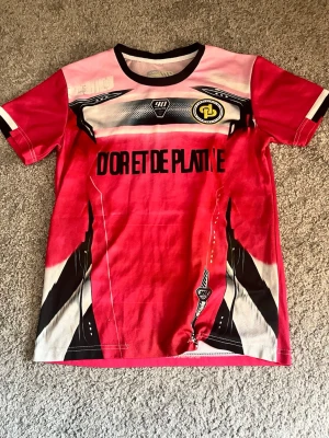 TOTO&NINETTA tröja - Cool röd t-shirt med svart och vit grafik, tryck fram och bak med texten 'D'OR ET DE PLATINE', 'Jul', 'Yamaha' och alienmotiv. Kortärmad modell med sportig känsla och svarta detaljer vid hals och ärmslut. Perfekt för dig som gillar unika statement-plagg.