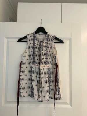 Vit blus odd Molly - Ärmlös blus i vitt med blått broderat mönster och dekorativa knappar framtill från Odd Molly. Blusen har smockad överdel och vinröda knytband i sidan som ger en snygg siluett. Textdetalj framtill och broderi bak i nacken. 