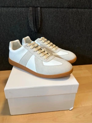 Vita och beige sneakers med gummisula - Stilrena sneakers i vitt och beige med ovandel i mocka och slät textil. Skorna har beige snörning och en klassisk brun gummisula med mönstrad undersida för bra grepp. Perfekta för en clean och avslappnad look.