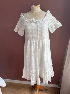 Vit sommarklänning med volang Odd Molly stl 2 - Söt vit klänning med korta ärmar, volangdetaljer och knyt framtill. Klänningen har en offshoulder-design och är tillverkad i ett lätt material, perfekt för varma dagar. Den har en romantisk känsla med rynkade detaljer och en luftig kjol.