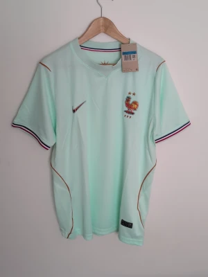 Ljusgrön Nike fotbollströja Frankrike 1 i S och 1 i M  - Snygg ljusgrön fotbollströja från Nike med Frankrikes landslagsmärke och två stjärnor på bröstet. Tröjan har korta ärmar, tunna bronsfärgade detaljer och randiga muddar i blått, vitt och rött. Perfekt för match eller träning.