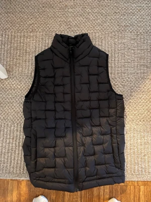 Everest väst - Snygg svart dunväst med quiltat mönster och hög krage. Västen har dragkedja framtill och är ärmlös, perfekt att slänga över en hoodie eller tröja för extra värme. Enkel och stilren design som passar till det mesta.